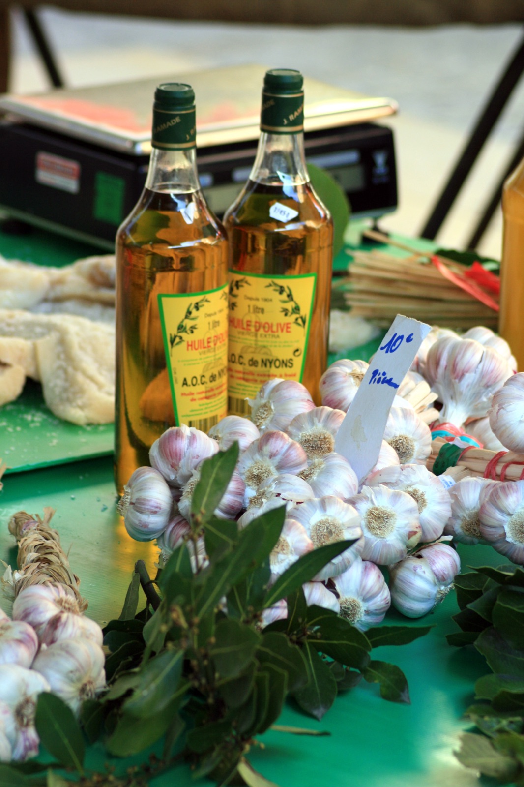 Deux bouteilles d’un litre d’huile d’olive dorée étiquetées vierge extra A.O.C. de Nyons sur un étal de marché, à côté de tresses d’ail, de feuilles de laurier et de pain.
