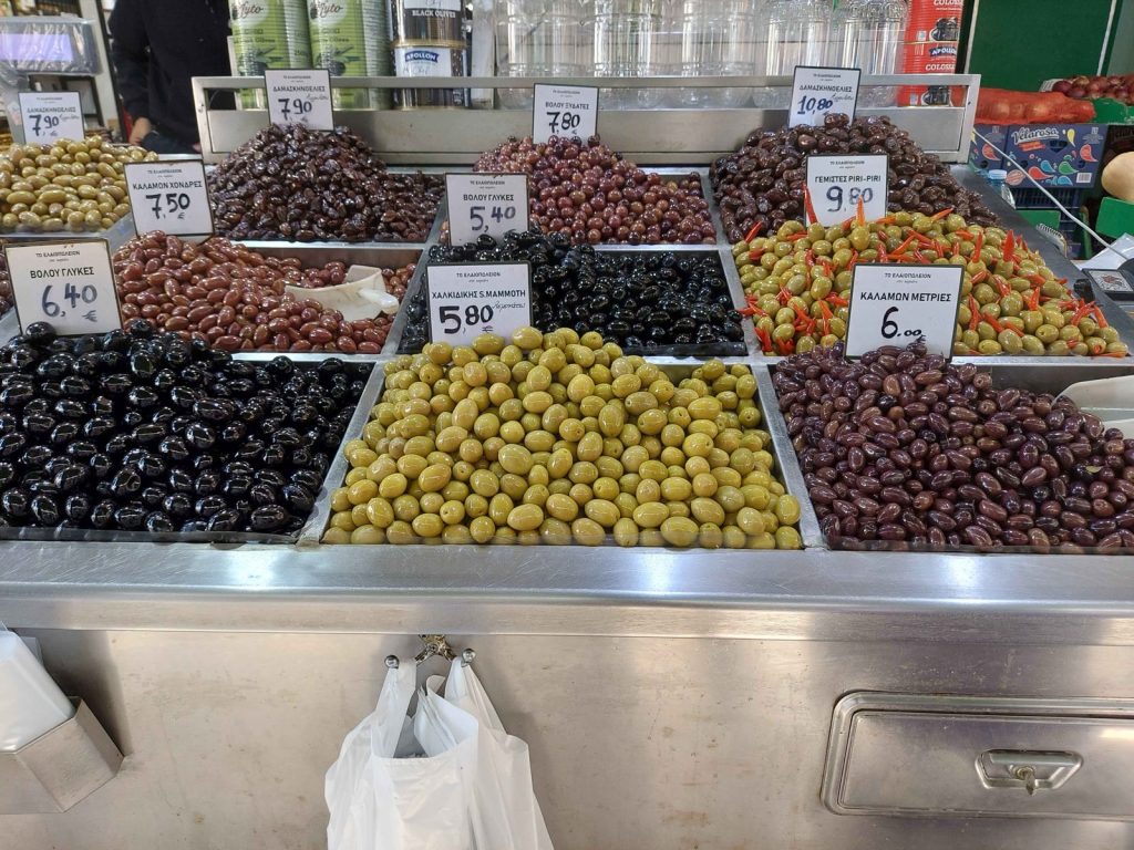 Retail olive counter at Kapani Market in Thessaloniki with about ten open bins of loose olives, each with a handwritten price per kilo in euros