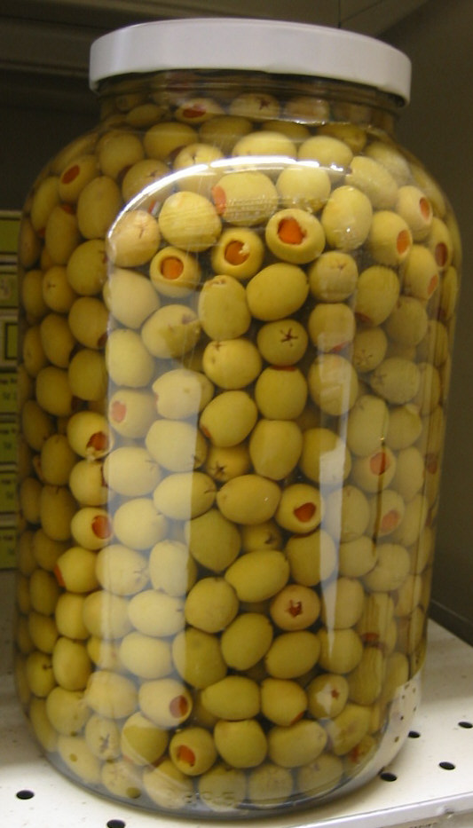 Grand bocal en verre à couvercle blanc rempli d'olives vertes farcies au piment, sur un rayon de magasin.