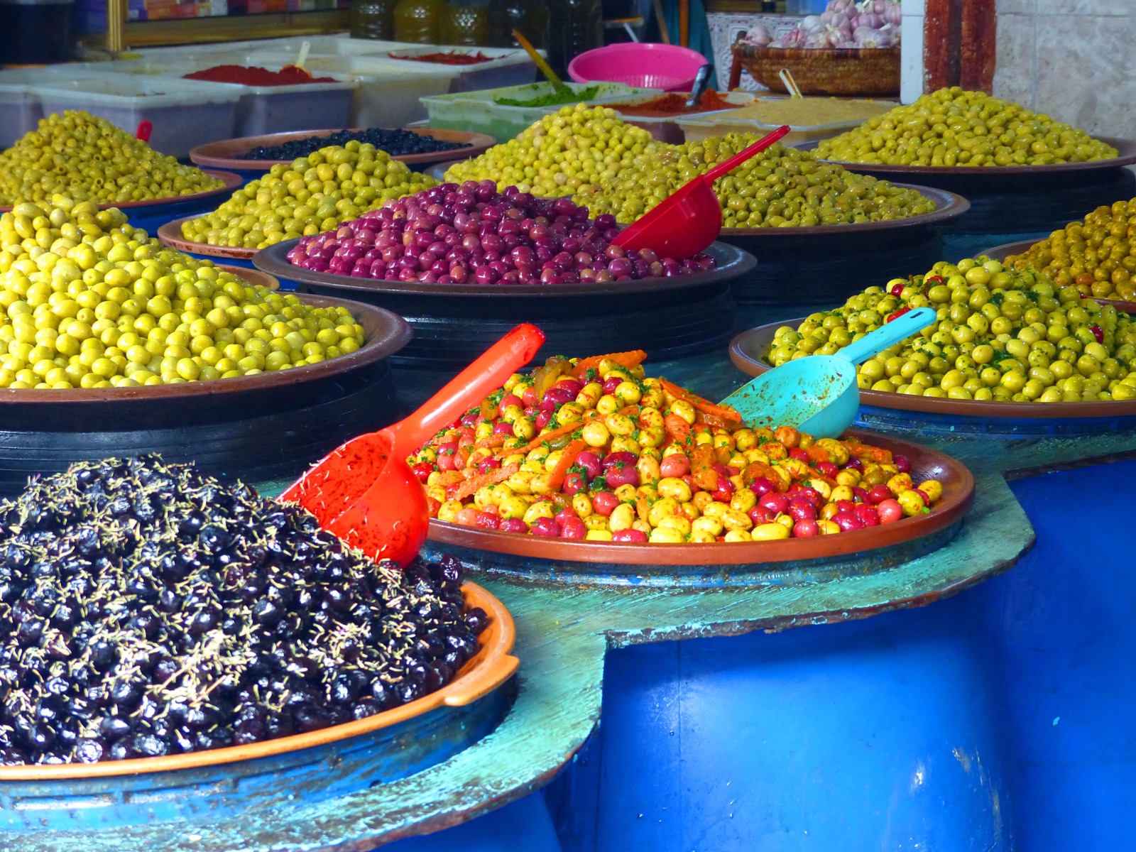 Wide ceramic platters heaped with green, violet, black and spiced marinated olives on a market stall counter, each with its own plastic scoop.
