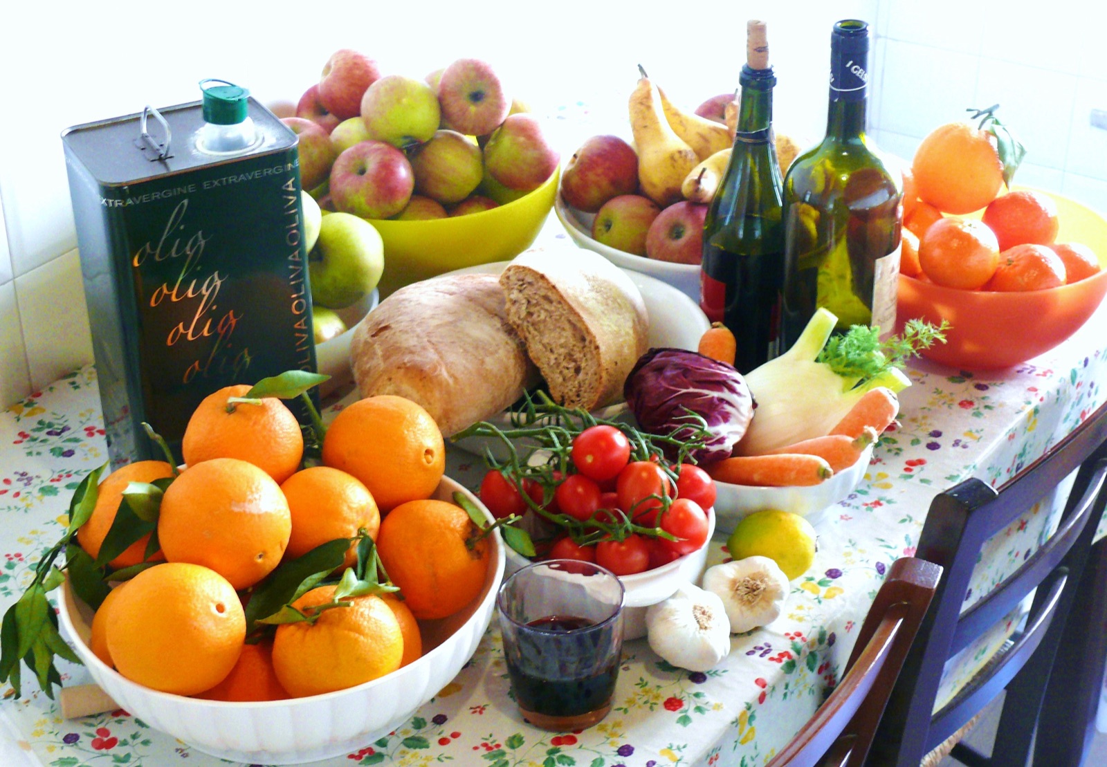 A kitchen table laid with a tin of extra virgin olive oil, bowls of oranges, apples and pears, cherry tomatoes on the vine, fennel, red cabbage, carrots, garlic and a rustic loaf.