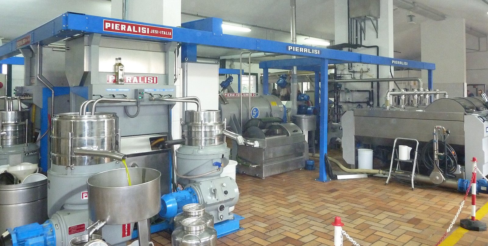 The interior of a modern olive mill: blue Pieralisi framework, stainless malaxers and horizontal decanter centrifuges, with fresh oil running into a steel tub.