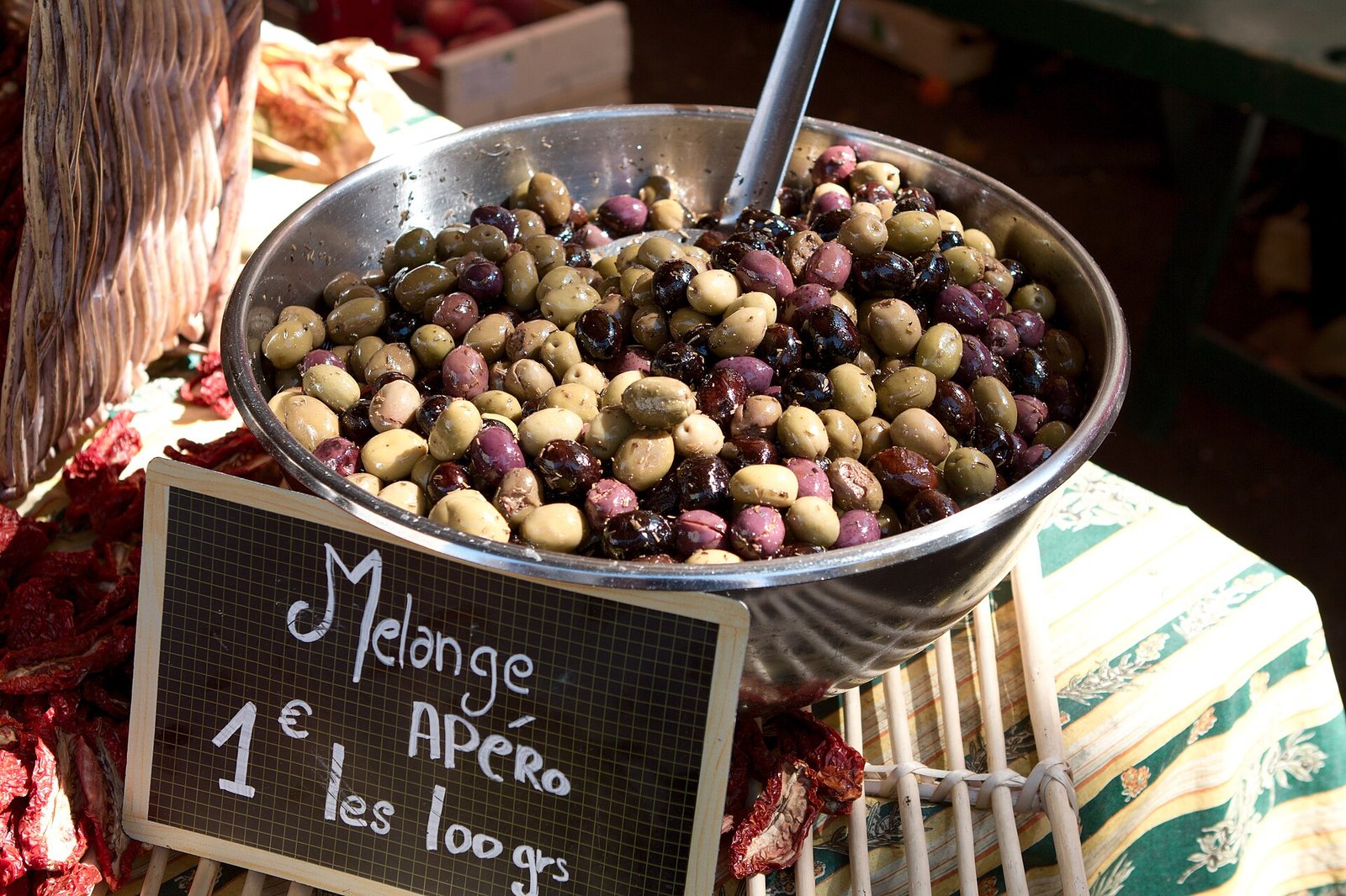 A large steel bowl of mixed marinated olives — green, tan, purple and black — on a Provençal market stall beside a chalkboard reading “Melange APÉRO”.