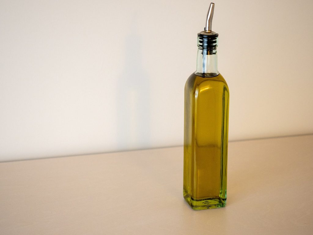A square glass cruet of golden-green olive oil fitted with a steel pouring spout, standing on a plain worktop.