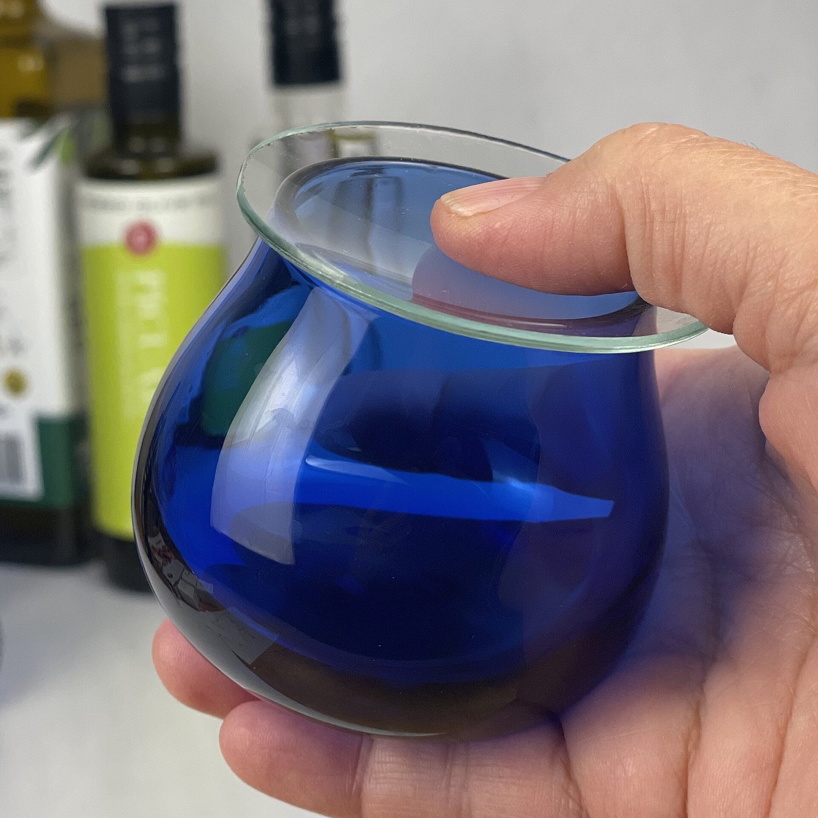 A hand holding the standard cobalt-blue olive oil tasting glass with its watch-glass cover, bottles of oil blurred behind.