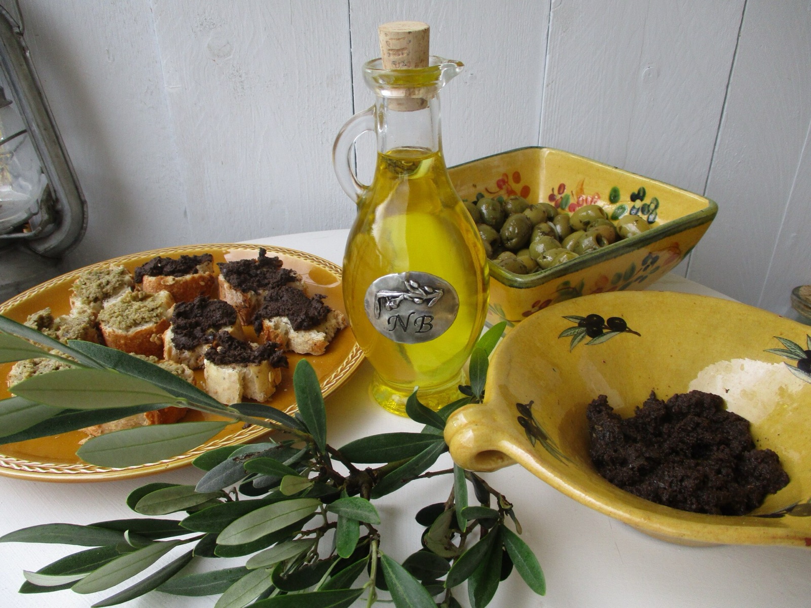 Un flacon en verre rempli d'huile d'olive dorée, entouré de plats provençaux : olives vertes, tapenade noire et tranches de pain grillé.